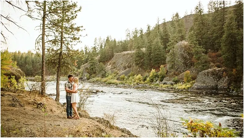 A couple embraces by a tree on a rocky Snohomish riverbank, surrounded by pine trees and greenery, with a flowing river and forested hills in the background. Snohomish Wedding Photography