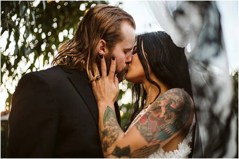 A couple kisses passionately outdoors in Snohomish; the man wears a black suit and has long hair, while the woman, in a white dress, has long dark hair and colorful tattoos. Sunlight filters through greenery in the background. Snohomish Wedding Photography