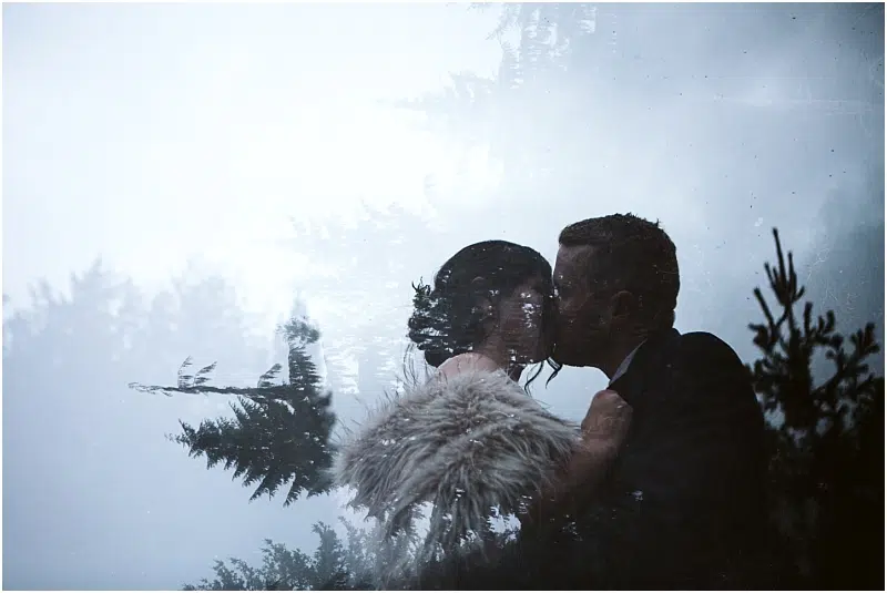 A couple dressed formally shares a kiss outdoors in Snohomish, surrounded by misty trees. The image has a dreamy, double-exposure effect, with tree branches blending into the couple's silhouettes. Snohomish Wedding Photography