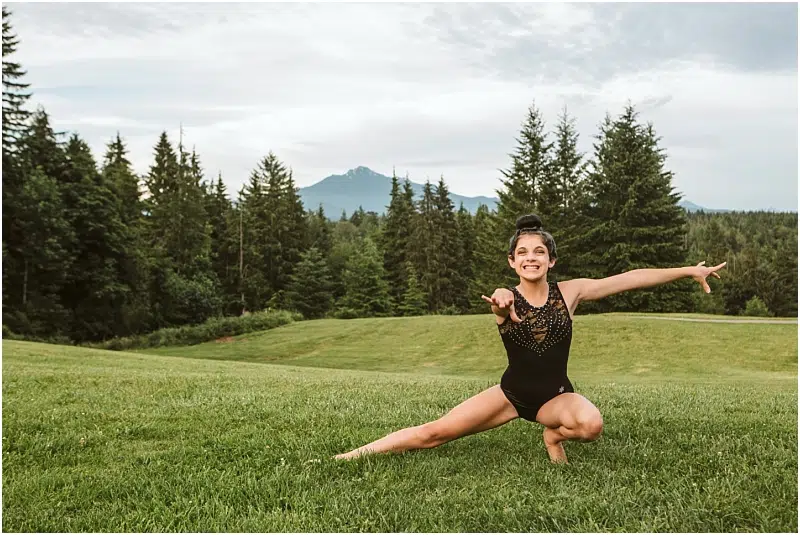 A young girl in a black leotard poses gracefully on one knee in a grassy Snohomish field, surrounded by tall evergreen trees and mountains under a partly cloudy sky. Snohomish Wedding Photography