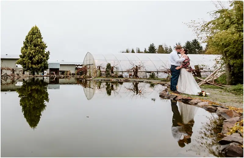 A bride and groom stand beside a small pond in Snohomish, sharing an embrace. Their reflection shimmers in the still water, with greenhouses and trees providing a picturesque backdrop on a cloudy day. Snohomish Wedding Photography
