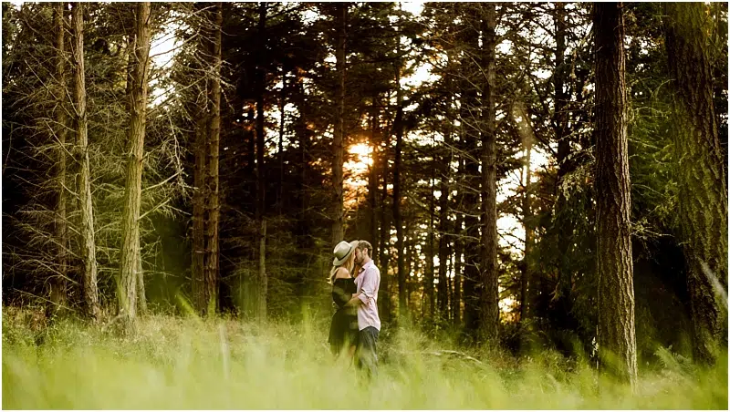 A couple stands close together in a Snohomish forest, kissing as sunlight filters through tall trees behind them. Grass in the foreground is blurred, adding depth to the romantic outdoor scene. Snohomish Wedding Photography