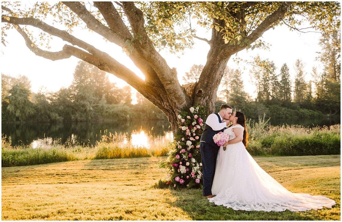 Summer Pemberton Farm Wedding with floral tree wrap and bride in white gown with bouquet kissing groom in blue suit in the golden sunset by the Snohomish River