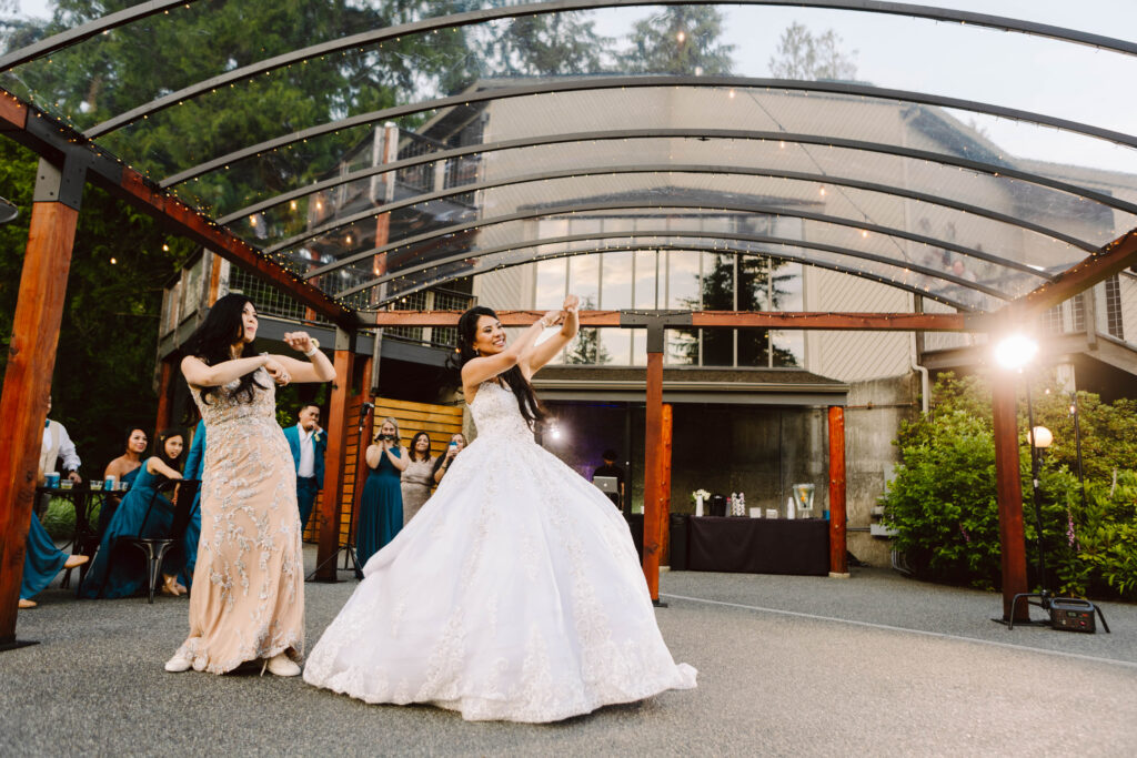 Snohomish Wedding Photographer GSquared Weddings Two women in elegant gowns dance under a transparent canopy with wooden beams. The woman in white, likely the bride, shares this joyous wedding moment with her mom. Guests in formal attire stand smiling and capturing photos at this lively outdoor celebration. Serving Seattle, Snohomish and Orlando