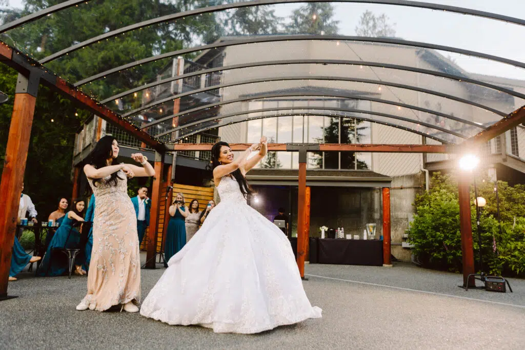 Snohomish Wedding Photographer GSquared Weddings Two women in elegant gowns dance under a transparent canopy with wooden beams. The woman in white, likely the bride, shares this joyous wedding moment with her mom. Guests in formal attire stand smiling and capturing photos at this lively outdoor celebration. Serving Seattle, Snohomish and Orlando