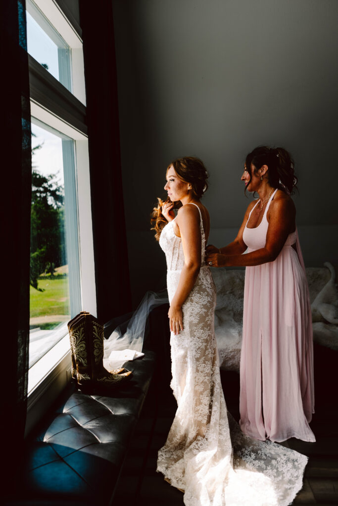Snohomish Wedding Photographer GSquared Weddings A bride in a lace gown stands near a window, her mother in a light pink dress lovingly adjusting her attire. Sunlight streams in, illuminating their figures. A pair of cowboy boots rests on the windowsill, honoring moms love for country flair at the wedding. Serving Seattle, Snohomish and Orlando