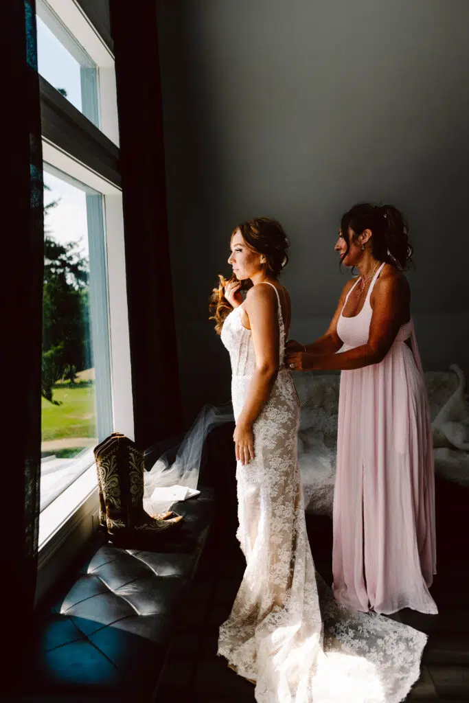 Snohomish Wedding Photographer GSquared Weddings A bride in a lace gown stands near a window, her mother in a light pink dress lovingly adjusting her attire. Sunlight streams in, illuminating their figures. A pair of cowboy boots rests on the windowsill, honoring moms love for country flair at the wedding. Serving Seattle, Snohomish and Orlando