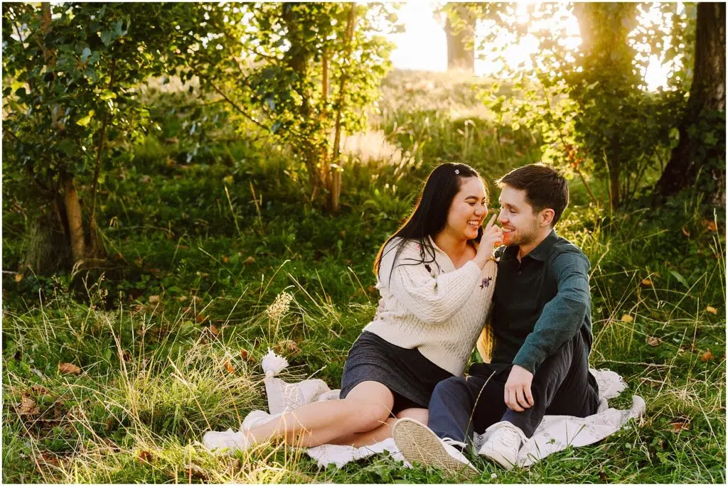 Snohomish Wedding Photographer GSquared Weddings A couple sits on a blanket in a sunlit forest clearing, smiling at each other with the joy of their newfound engagement. The woman wears a white sweater and the man a dark green shirt, surrounded by grass and trees as sunlight filters through the leaves. Serving Seattle, Snohomish and Orlando