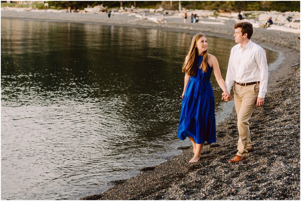 Snohomish Wedding Photographer GSquared Weddings A couple walks hand in hand along a pebbled beach. The woman wears a blue dress, and the man wears a white shirt and beige pants. The calm water reflects the evening light, and driftwood is scattered along the shore. Serving Seattle, Snohomish and Orlando