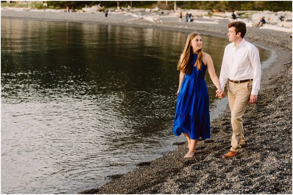 Snohomish Wedding Photographer GSquared Weddings A couple walks hand in hand along a pebbled beach. The woman wears a blue dress, and the man wears a white shirt and beige pants. The calm water reflects the evening light, and driftwood is scattered along the shore. Serving Seattle, Snohomish and Orlando