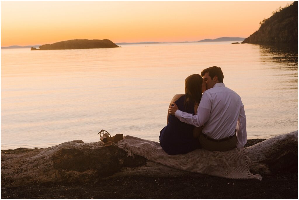 Snohomish Wedding Photographer GSquared Weddings A couple sits on a blanket at a beach during sunset, embracing as they look at the calm sea. A pair of sandals is placed nearby, and rocky islands are visible in the distance against the warm, glowing sky. Serving Seattle, Snohomish and Orlando