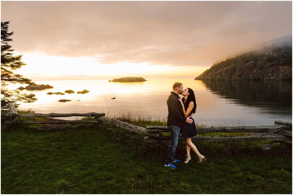 Snohomish Wedding Photographer GSquared Weddings A couple embraces and kisses by a wooden fence overlooking a serene lake at sunset. The sky is colorful, with clouds and a glowing horizon. Rocky hills and small islands are visible in the background, reflecting on the calm water. Serving Seattle, Snohomish and Orlando