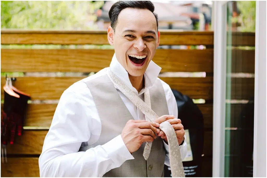 Snohomish Wedding Photographer GSquared Weddings A man in a gray vest and white shirt laughs while adjusting his tie. The background features a wooden fence and some greenery. Serving Seattle, Snohomish and Orlando