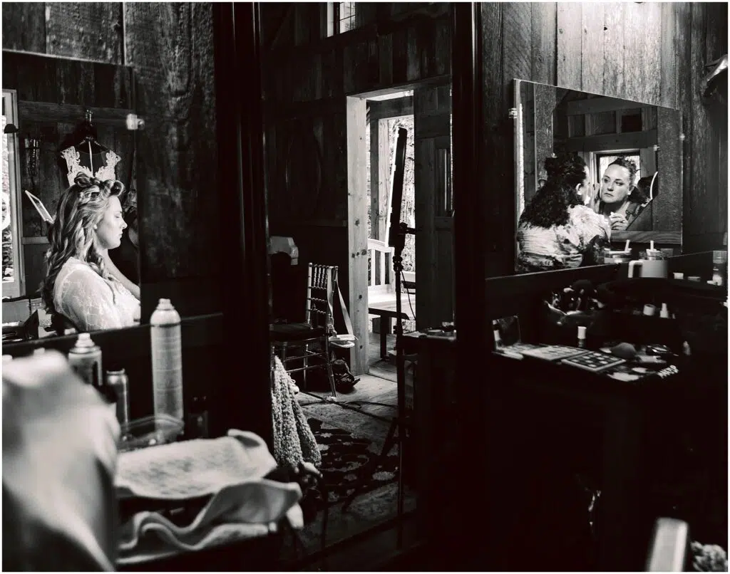 Snohomish Wedding Photographer GSquared Weddings A woman is preparing for a wedding, sitting in a wooden room. She is wearing a robe and tiara, looking at herself in a mirror. Wedding accessories and makeup items are on the table nearby. Theres a doorway leading to an outside view. Serving Seattle, Snohomish and Orlando