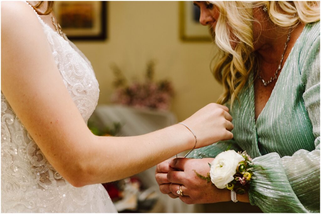 Snohomish Wedding Photographer GSquared Weddings A bride in a white lace dress assists a woman in a green dress with a white rose corsage. The moment captures the intimacy and elegance of a special occasion, likely a wedding. Floral decor is visible in the background. Serving Seattle, Snohomish and Orlando