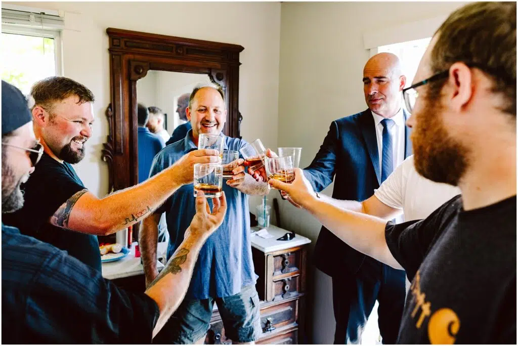 Snohomish Wedding Photographer GSquared Weddings A group of six men stand in a room, raising glasses in a celebratory toast. They are smiling and appear to be in a festive mood, with a mirror and wooden furniture in the background. Serving Seattle, Snohomish and Orlando