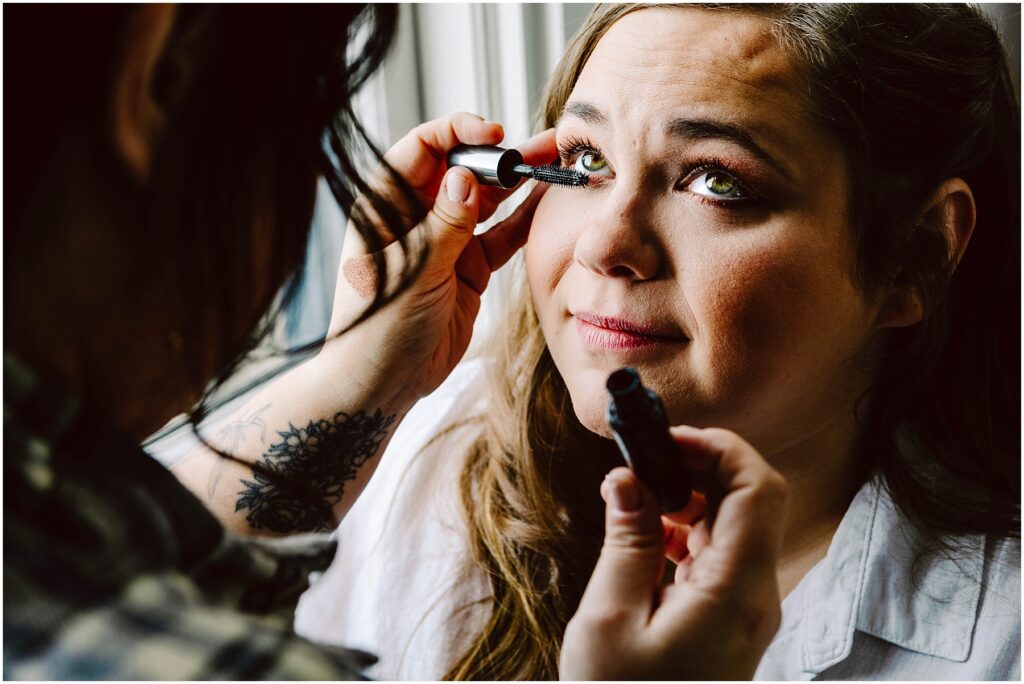Snohomish Wedding Photographer GSquared Weddings A woman with long brown hair is having mascara applied to her eyelashes by another person. She is wearing a white shirt and sitting by a window, looking slightly upward with a gentle expression. Serving Seattle, Snohomish and Orlando