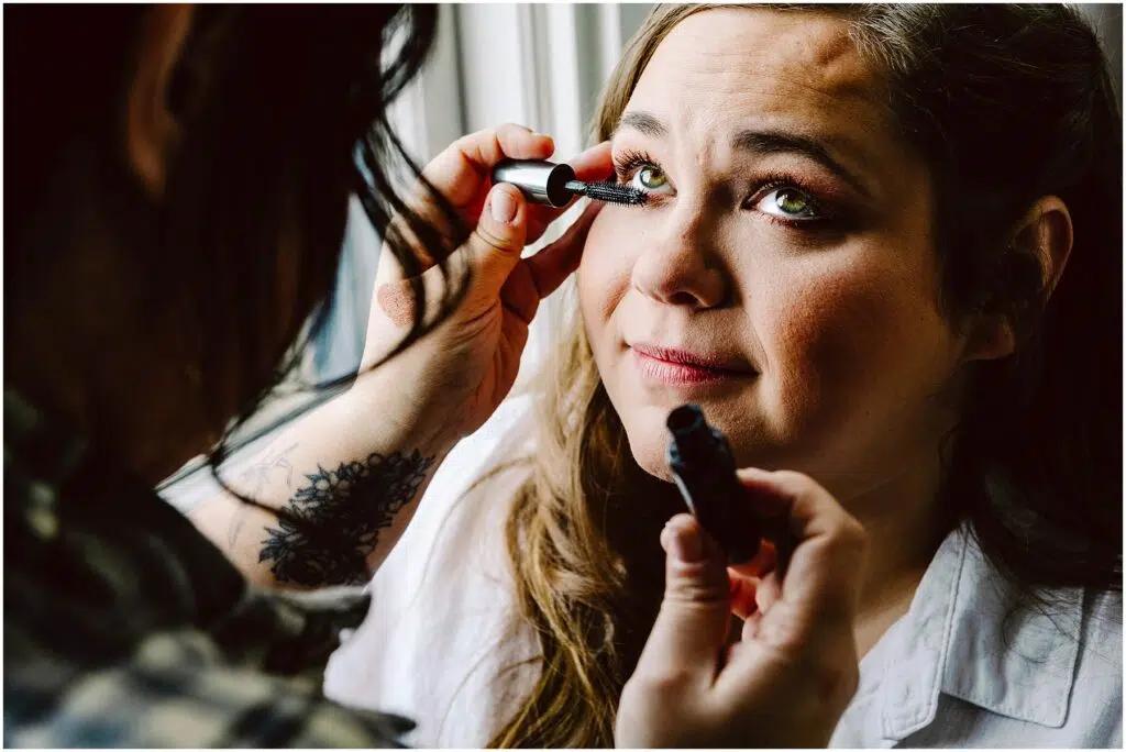 Snohomish Wedding Photographer GSquared Weddings A woman with long brown hair is having mascara applied to her eyelashes by another person. She is wearing a white shirt and sitting by a window, looking slightly upward with a gentle expression. Serving Seattle, Snohomish and Orlando