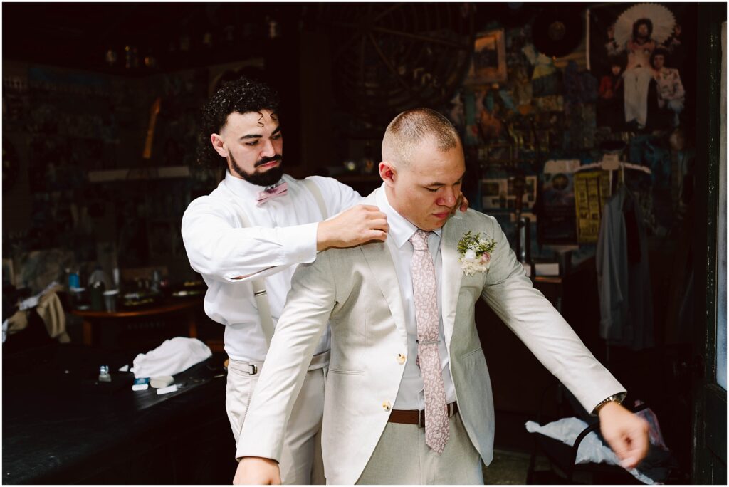 Snohomish Wedding Photographer GSquared Weddings A groom in a beige suit is being helped with his jacket by a groomsman wearing a white outfit and pink bow tie. They are standing in a room with eclectic decorations and artwork on the walls. Serving Seattle, Snohomish and Orlando