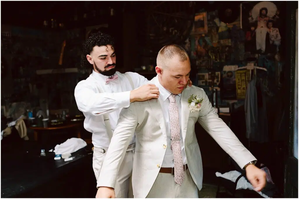 Snohomish Wedding Photographer GSquared Weddings A groom in a beige suit is being helped with his jacket by a groomsman wearing a white outfit and pink bow tie. They are standing in a room with eclectic decorations and artwork on the walls. Serving Seattle, Snohomish and Orlando
