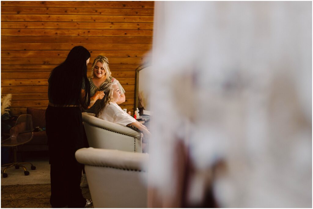 Snohomish Wedding Photographer GSquared Weddings Two women style another womans hair and makeup in a salon with wooden walls. The seated woman is smiling while two stylists work on her. Blurred foreground elements frame the scene, adding depth to the cozy setting. Serving Seattle, Snohomish and Orlando