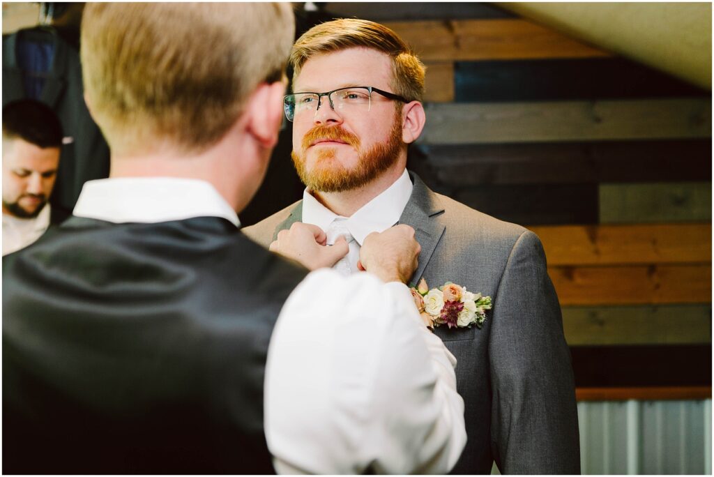 Snohomish Wedding Photographer GSquared Weddings A groom with a beard stands facing forward, wearing a gray suit and a floral boutonniere. Another person, seen from behind, adjusts the grooms bow tie. In the background, wood panels and another person are visible. Serving Seattle, Snohomish and Orlando