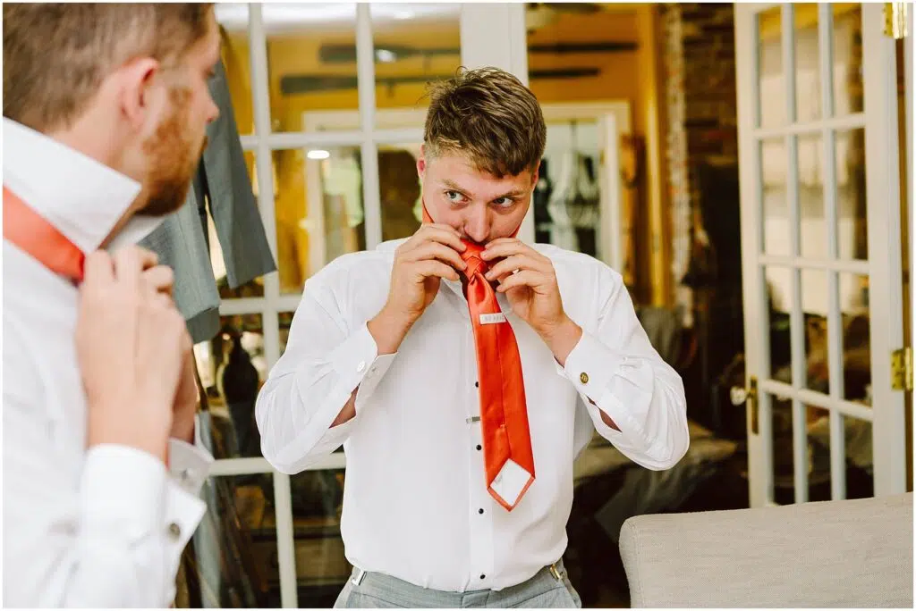 Snohomish Wedding Photographer GSquared Weddings A person in a white shirt, reflected in a mirror, bites open the packaging of an orange tie in a room with glass-paneled doors and a suit hanging in the background. Serving Seattle, Snohomish and Orlando