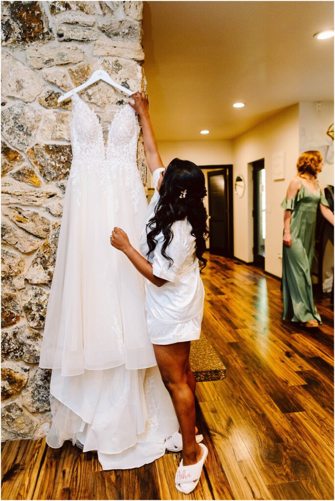 Snohomish Wedding Photographer GSquared Weddings A woman with long dark hair, wearing a white robe and slippers, reaches toward a white wedding dress hanging on a stone wall. The room has wooden floors and another person is seen in the background wearing a green dress. Serving Seattle, Snohomish and Orlando