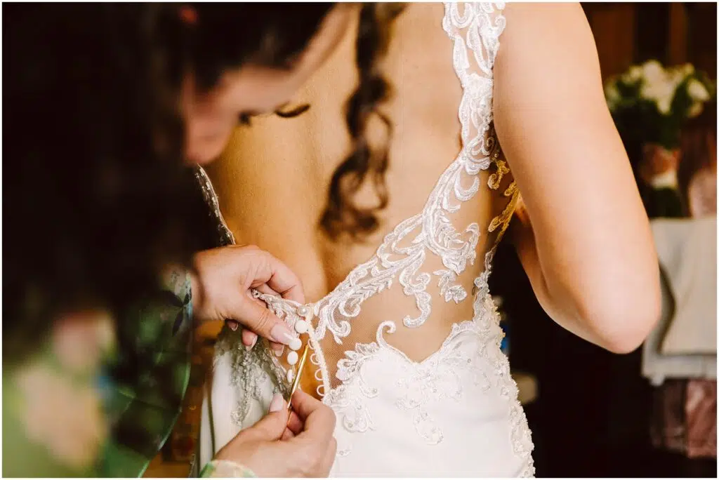 Snohomish Wedding Photographer GSquared Weddings A close-up of a bride in a lace wedding dress being buttoned up by an assistant. Her back is facing the camera, revealing intricate lace details on the dress. Serving Seattle, Snohomish and Orlando