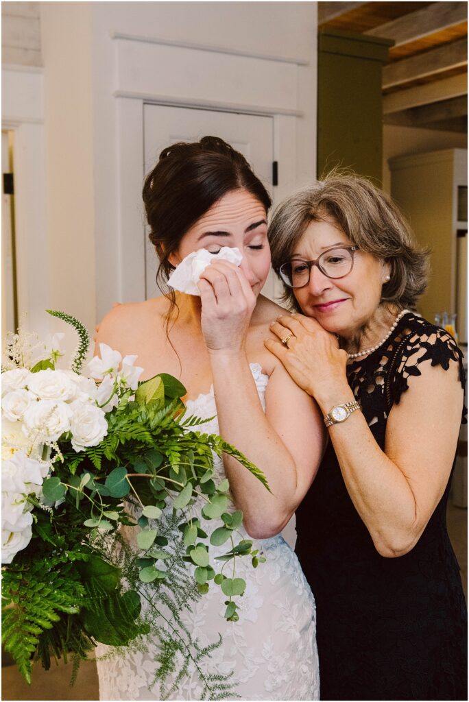 Snohomish Wedding Photographer GSquared Weddings A bride in a white dress holds a large bouquet of white flowers and greenery, wiping tears from her eyes. An older woman in a black dress, likely a family member, stands close, affectionately resting her head on the brides shoulder. Serving Seattle, Snohomish and Orlando