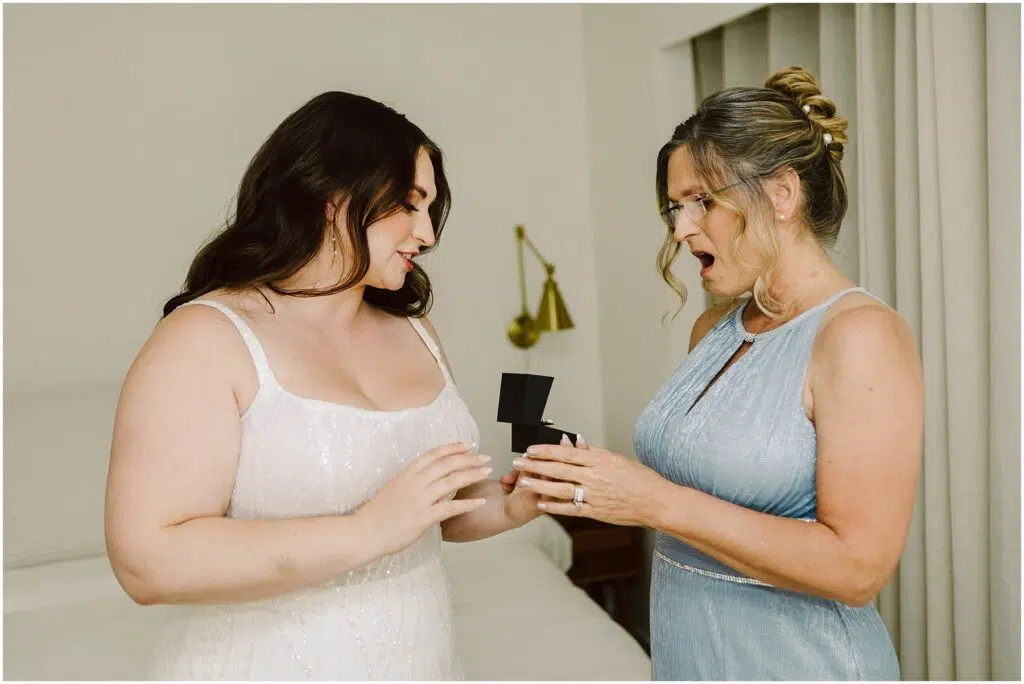 Snohomish Wedding Photographer GSquared Weddings Two women stand in a room, one in a white dress holding an open black box; the other in a light blue dress looks surprised and joyful. They appear to be celebrating a special occasion. A bed and a wall lamp are in the background. Serving Seattle, Snohomish and Orlando