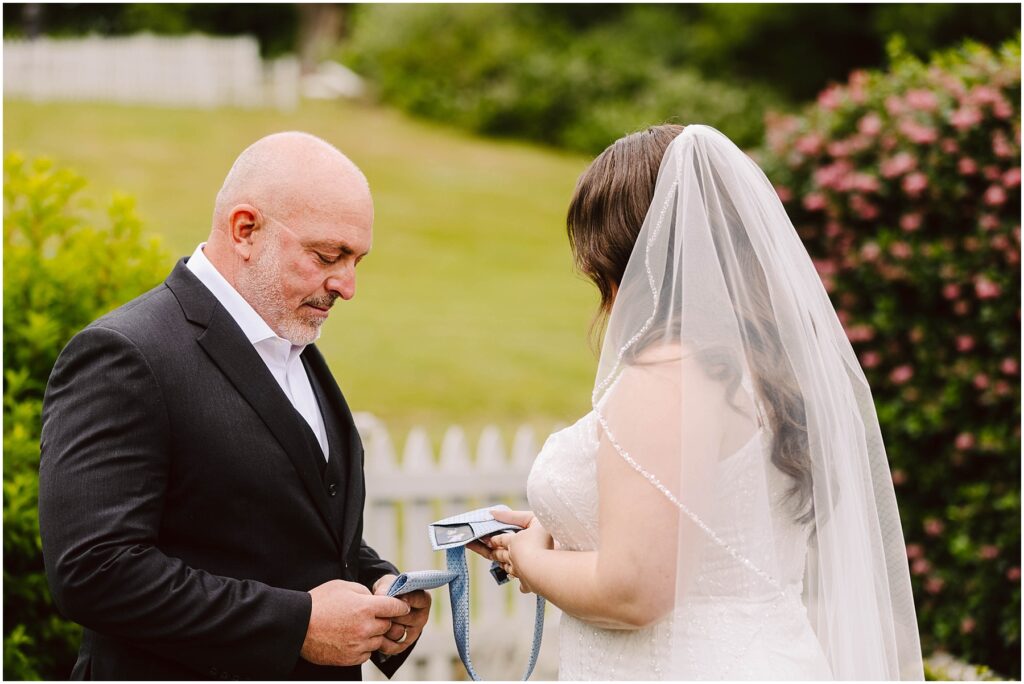 Snohomish Wedding Photographer GSquared Weddings A bride and a man in a suit, possibly her father, stand outdoors in a garden setting. The bride, facing away, holds a piece of paper. The man looks down at a tie in his hands. A white fence and greenery are in the background. Serving Seattle, Snohomish and Orlando