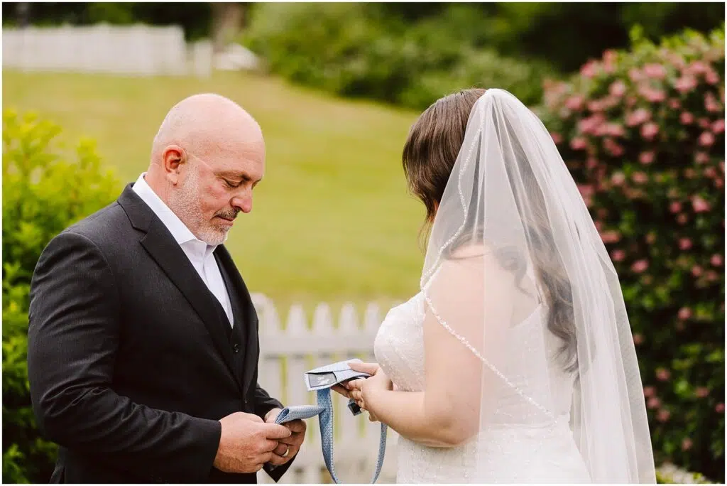 Snohomish Wedding Photographer GSquared Weddings A bride and a man in a suit, possibly her father, stand outdoors in a garden setting. The bride, facing away, holds a piece of paper. The man looks down at a tie in his hands. A white fence and greenery are in the background. Serving Seattle, Snohomish and Orlando
