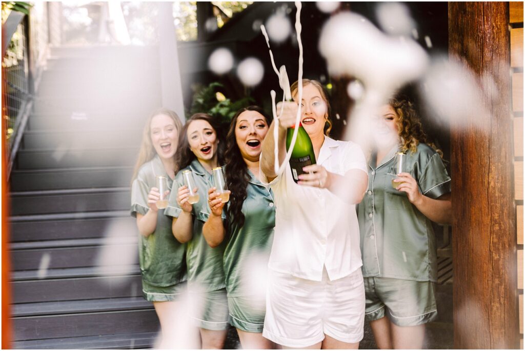 Snohomish Wedding Photographer GSquared Weddings A group of five women in matching pajamas, one in white and the others in green, are excitedly popping a bottle of champagne outdoors. The cork flies and champagne sprays into the air, capturing a moment of joyful celebration. Serving Seattle, Snohomish and Orlando