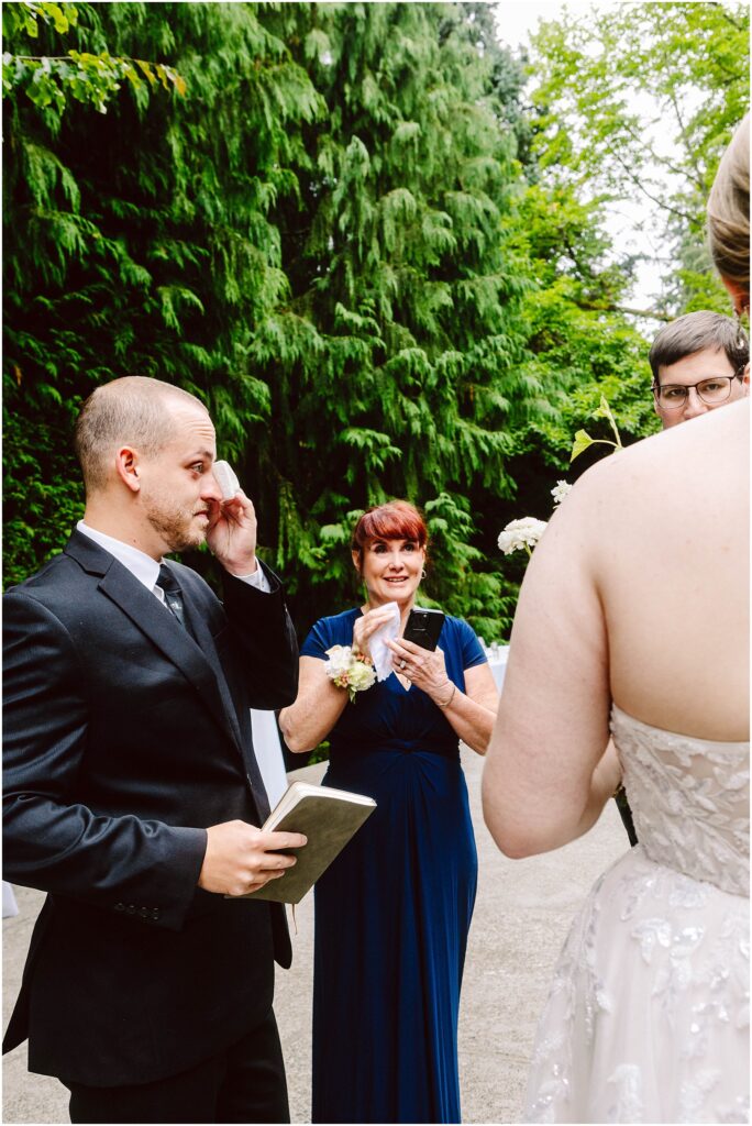Snohomish Wedding Photographer GSquared Weddings A groom in a suit stands outdoors, holding a book and wiping a tear. A woman in a blue dress, smiling, holds a smartphone. The bride, in a white dress, is partially visible. Lush green foliage surrounds them. Serving Seattle, Snohomish and Orlando