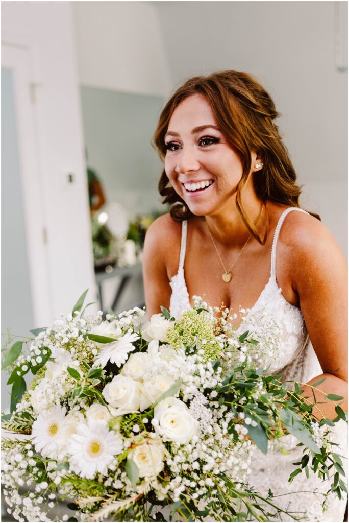 Snohomish Wedding Photographer GSquared Weddings A smiling bride with long brown hair holds a large bouquet of white flowers and greenery. She is wearing a white lace dress with thin straps and a gold necklace. The background is softly blurred, focusing on the brides joyful expression. Serving Seattle, Snohomish and Orlando