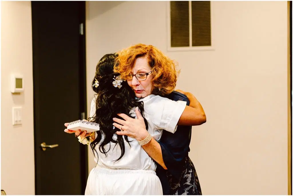 Snohomish Wedding Photographer GSquared Weddings A woman with curly red hair hugs another woman with long black hair and a floral hair accessory. The woman being hugged holds a small, white, rectangular object. They are standing in a room with beige walls and a black door. Serving Seattle, Snohomish and Orlando