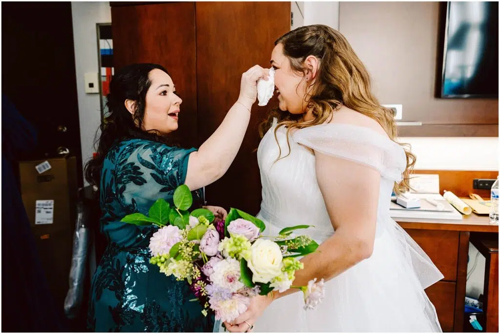 Snohomish Wedding Photographer GSquared Weddings A woman in a white wedding dress holds a bouquet of flowers while another woman, in a teal dress, gently dabs her face with a tissue. They are in a room with wooden cabinets and soft lighting. Serving Seattle, Snohomish and Orlando