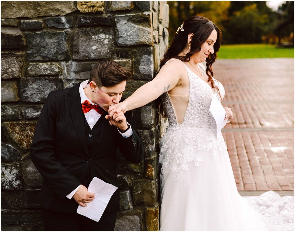 Snohomish Wedding Photographer GSquared Weddings A person in a suit and red bow tie kisses the hand of a person in a white wedding dress. Both hold pieces of paper and stand near a stone wall, outdoors on a brick pathway. Serving Seattle, Snohomish and Orlando