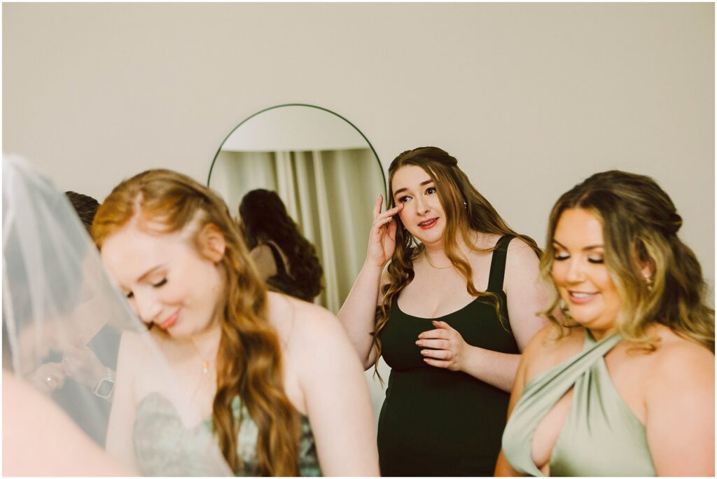 Snohomish Wedding Photographer GSquared Weddings Three women in elegant dresses are in a softly lit room, two of them smiling and looking down, while one wipes her eye gently. A round mirror is in the background, reflecting part of the scene. Serving Seattle, Snohomish and Orlando