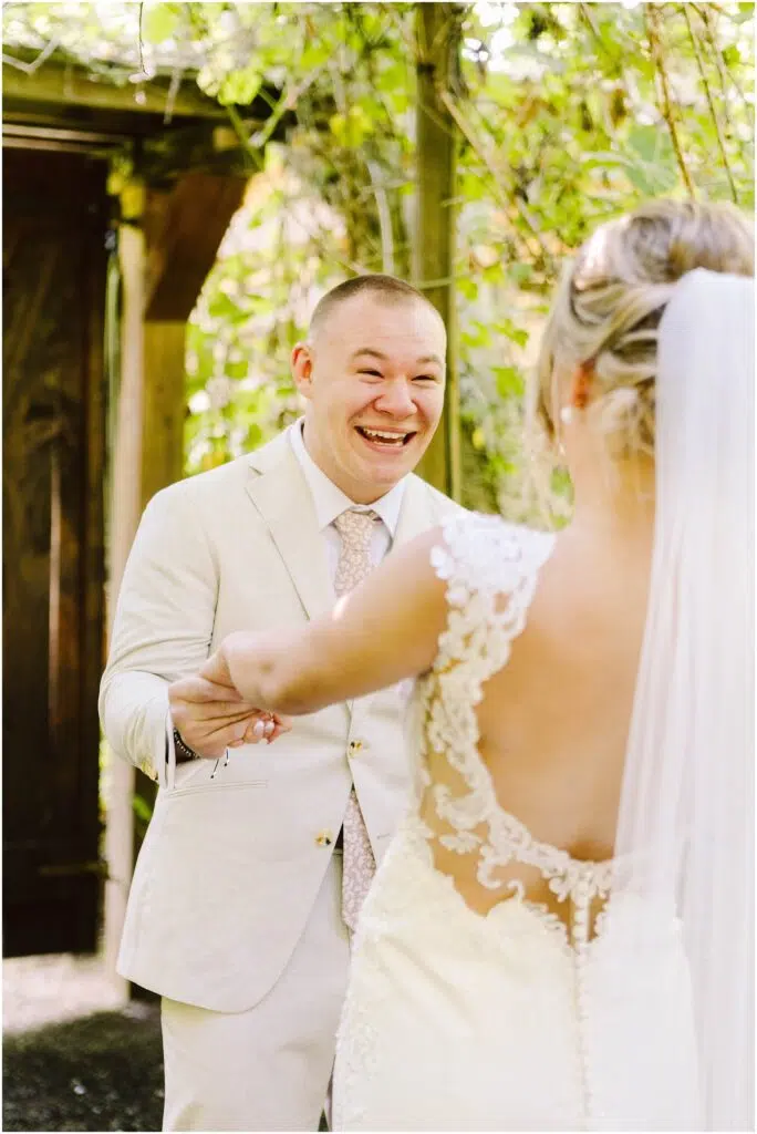 Snohomish Wedding Photographer GSquared Weddings Groom in a light beige suit smiling joyfully, holding hands with a bride in a lace dress with a veil, facing him. They are outdoors, surrounded by green foliage and wooden structures. Serving Seattle, Snohomish and Orlando