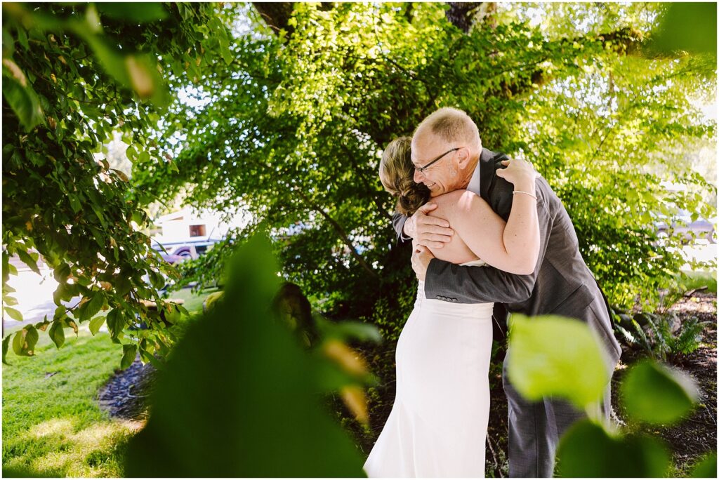 Snohomish Wedding Photographer GSquared Weddings A bride in a white dress hugs an older man in a suit amidst lush greenery. Sunlight filters through the leaves, casting soft light on the scene. The foliage frames the couple, creating an intimate, joyful moment. Serving Seattle, Snohomish and Orlando
