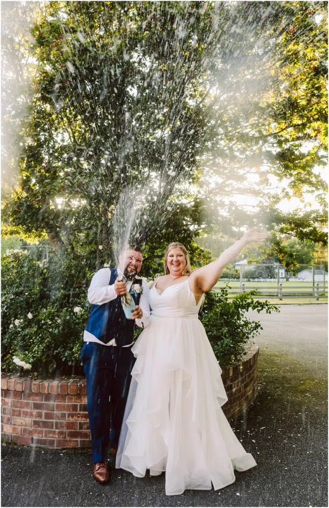 Snohomish Wedding Photographer GSquared Weddings A bride and groom celebrate by popping a champagne bottle, with the cork flying and champagne spraying. They stand in front of a lush tree, with sunlight filtering through the leaves. The bride smiles and raises her arm joyfully. Serving Seattle, Snohomish and Orlando