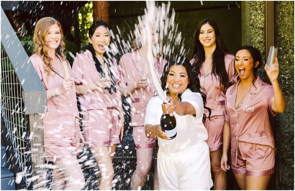Snohomish Wedding Photographer GSquared Weddings A woman in white pajamas joyfully pops open a champagne bottle, spraying it around her. Five women in matching pink pajamas laugh and react with excitement. They are outside, with greenery and a wooden structure in the background. Serving Seattle, Snohomish and Orlando