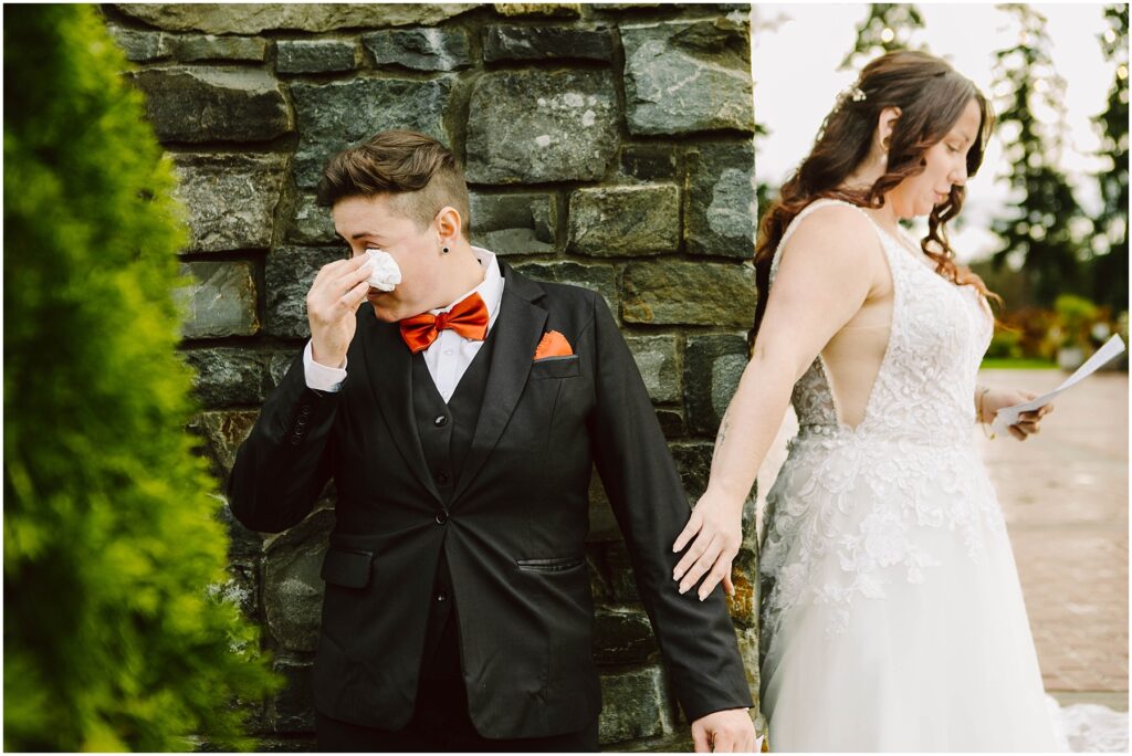 Snohomish Wedding Photographer GSquared Weddings A couple stands near a stone wall, both in formal attire. The person in a suit with a red bow tie wipes tears with a tissue. The other, in a white lace gown, gently touches their partners hand, holding a paper. Emotional moment in a wedding setting. Serving Seattle, Snohomish and Orlando