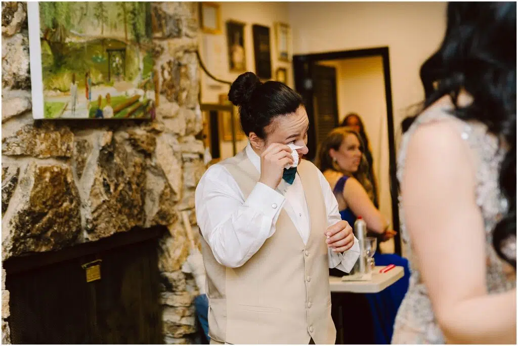 Snohomish Wedding Photographer GSquared Weddings A person in formal attire wipes away tears while standing near a stone fireplace. They wear a cream vest and a blue bow tie. A person in blue is seated in the background, and a framed painting hangs on the wall. Serving Seattle, Snohomish and Orlando