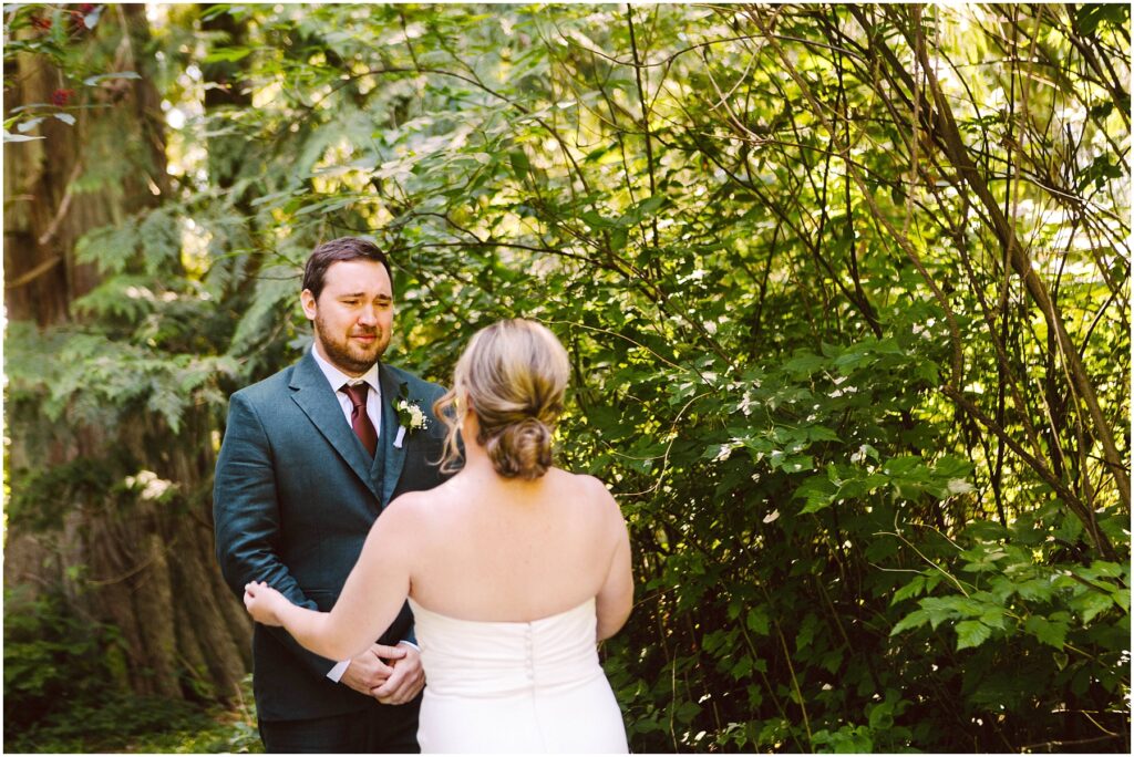 Snohomish Wedding Photographer GSquared Weddings A couple stands facing each other outdoors, surrounded by lush greenery. The groom, in a dark suit, looks at the bride, who is wearing a white dress with her hair up. Sunlight filters through the trees, creating a serene atmosphere. Serving Seattle, Snohomish and Orlando