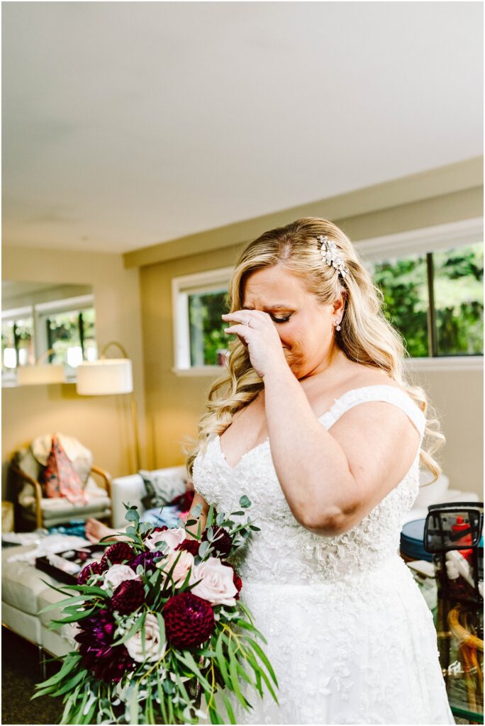 Snohomish Wedding Photographer GSquared Weddings A bride in a white lace gown and floral hairpiece wipes tears from her eyes, holding a bouquet with dark red and white flowers. She stands in a brightly lit room with modern furniture and large windows. Serving Seattle, Snohomish and Orlando
