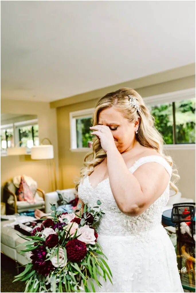 Snohomish Wedding Photographer GSquared Weddings A bride in a white lace gown and floral hairpiece wipes tears from her eyes, holding a bouquet with dark red and white flowers. She stands in a brightly lit room with modern furniture and large windows. Serving Seattle, Snohomish and Orlando