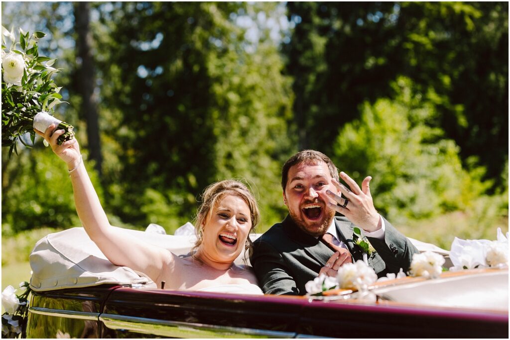 Snohomish Wedding Photographer GSquared Weddings A joyful bride and groom are sitting in a classic convertible. The bride holds a bouquet aloft and the groom shows off his wedding ring. Both are smiling widely against a backdrop of lush green trees. Serving Seattle, Snohomish and Orlando